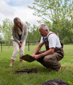 Guy crouching near septic tank point to clipboard with woman standing beside him